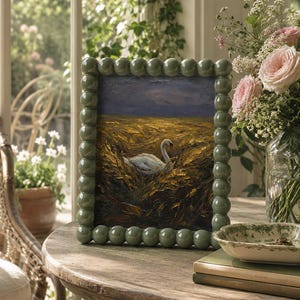 May include: A painting of a swan in a field, framed in a light green beaded frame. The painting sits on a wooden table, with a vase of pink roses and other flowers. A small dish and books are also on the table.