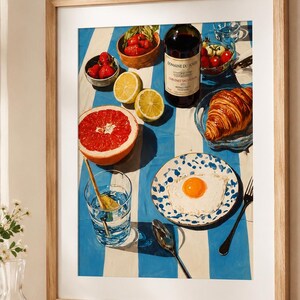 May include: A framed print depicting a breakfast setting. The image shows a bottle of wine, a croissant, a plate with a fried egg, strawberries, lemons, and a grapefruit on a blue and white striped tablecloth. A glass of water with a spoon is also visible.