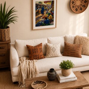 May include: A living room with a white sofa and cushions in brown and cream tones. A framed artwork of a market scene is above the sofa. A wooden coffee table holds a plant, books, and a decorative bowl, creating a warm, inviting atmosphere.