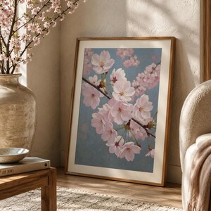 以下が含まれることがあります： 青い背景にピンク色の桜の花の枝を描いた額入りのアートプリント。桜の花の入った花瓶と木製のテーブルがある部屋に飾られています。 フレームは木製です。