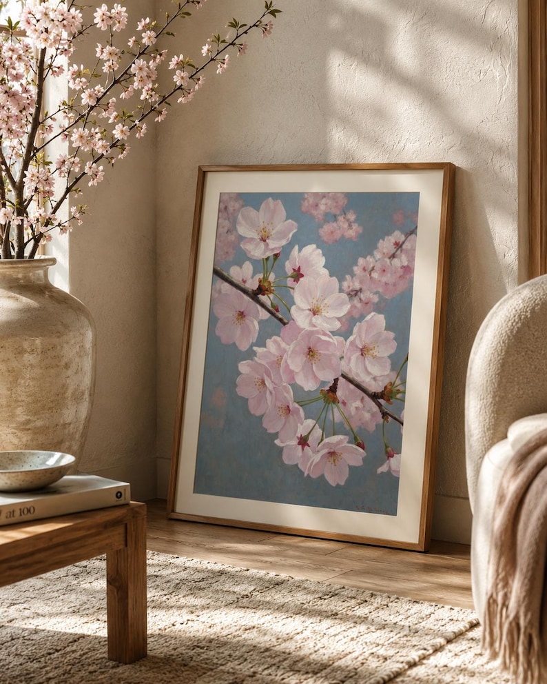 以下が含まれることがあります： 青い背景にピンク色の桜の花の枝を描いた額入りのアートプリント。桜の花の入った花瓶と木製のテーブルがある部屋に飾られています。 フレームは木製です。