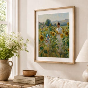 Puede incluir: Una impresi&oacute;n enmarcada de una pintura que representa a una mujer y un ni&ntilde;o en un campo de girasoles. La obra de arte est&aacute; en un marco de madera y cuelga de una pared beige. Un jarr&oacute;n con flores blancas y libros est&aacute;n sobre una mesa de madera debajo.