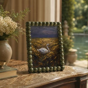 May include: A framed painting depicting a swan in a field, set against a dark blue sky. The frame is green and beaded. The artwork sits on a marble-topped wooden table. A vase of flowers and a book are also visible in the background.