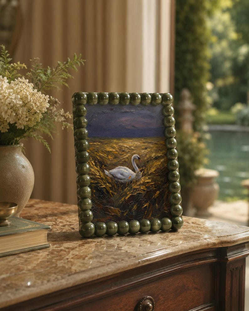 May include: A framed painting depicting a swan in a field, set against a dark blue sky. The frame is green and beaded. The artwork sits on a marble-topped wooden table. A vase of flowers and a book are also visible in the background.