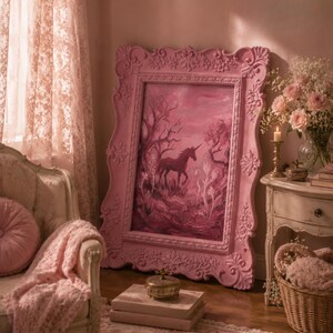 May include: A pink framed painting of a unicorn in a forest setting. The ornate frame is a light pink color. The painting is displayed in a room with a matching color scheme, including a chair, blanket, and curtains.