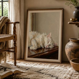 May include: A framed print of a sleeping white cat. The cat is resting on a dark surface, its eyes closed. The artwork is in a light wooden frame, leaning against a wall. The room has a neutral colour palette with a rug and chair.
