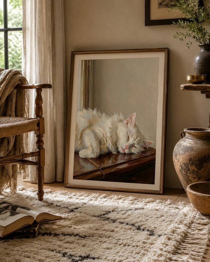 May include: A framed print of a sleeping white cat. The cat is resting on a dark surface, its eyes closed. The artwork is in a light wooden frame, leaning against a wall. The room has a neutral colour palette with a rug and chair.