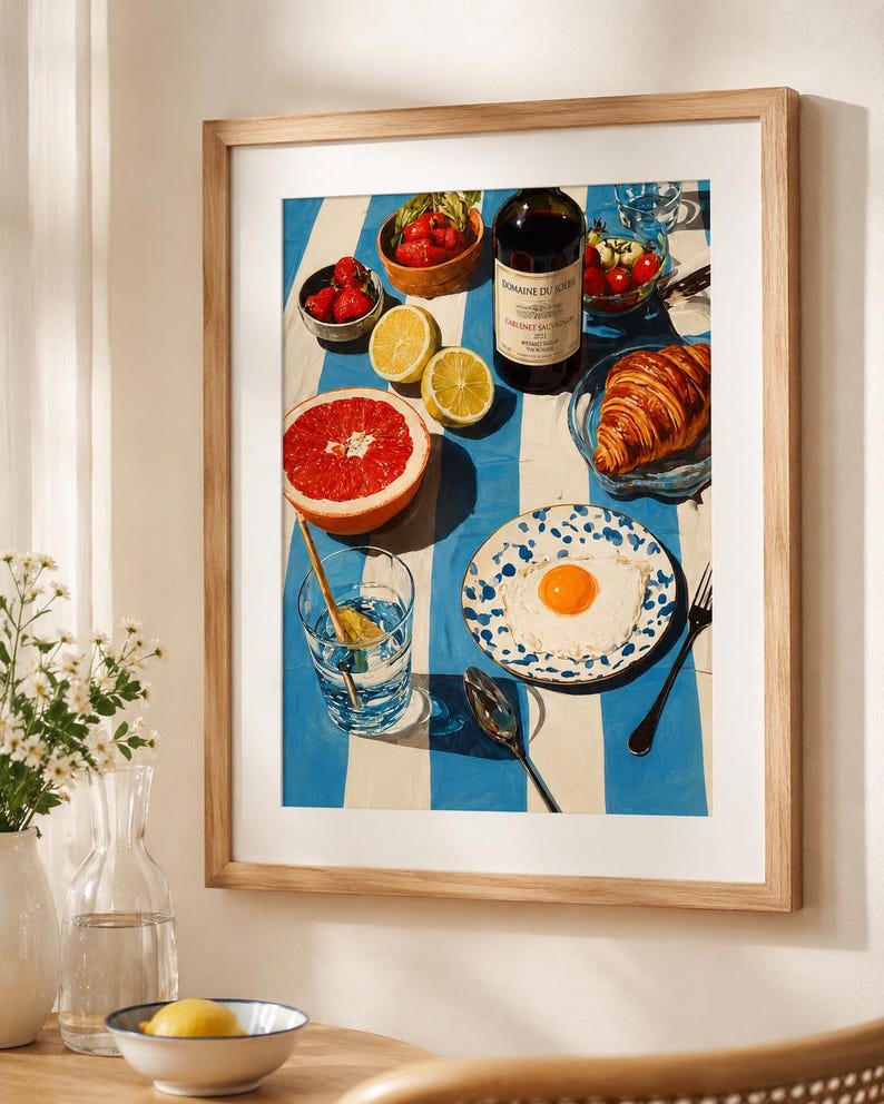 May include: A framed print depicting a breakfast setting. The image shows a bottle of wine, a croissant, a plate with a fried egg, strawberries, lemons, and a grapefruit on a blue and white striped tablecloth. A glass of water with a spoon is also visible.