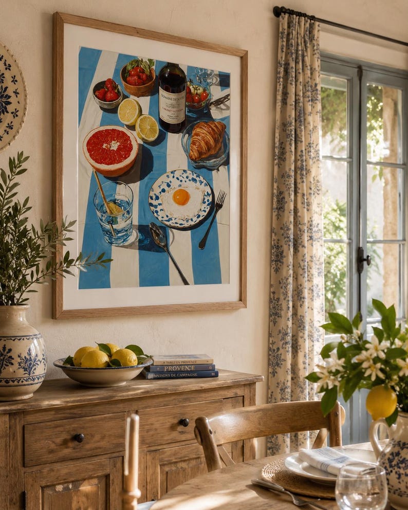 May include: A framed print shows a breakfast setting with a blue and white striped cloth. It includes a wine bottle, croissant, grapefruit, strawberries, lemons, and a fried egg on a plate. The print hangs above a wooden cabinet, with a bowl of lemons.