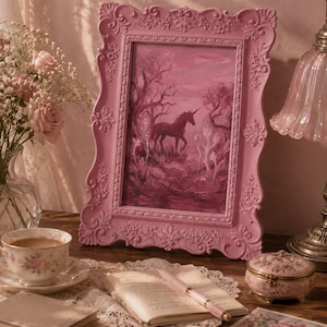 May include: A pink framed painting of a unicorn in a forest scene. The frame has ornate detailing. The painting is on a wooden table with a teacup, books, a small trinket box, and a pen.