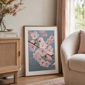以下が含まれることがあります： 淡いピンク色の桜の花が咲く枝が描かれた、青色の背景の額入りアートプリント。木製キャビネットとクリーム色の肘掛け椅子が置かれたリビングルームに飾られています。プリントは木製のフレームに入っています。