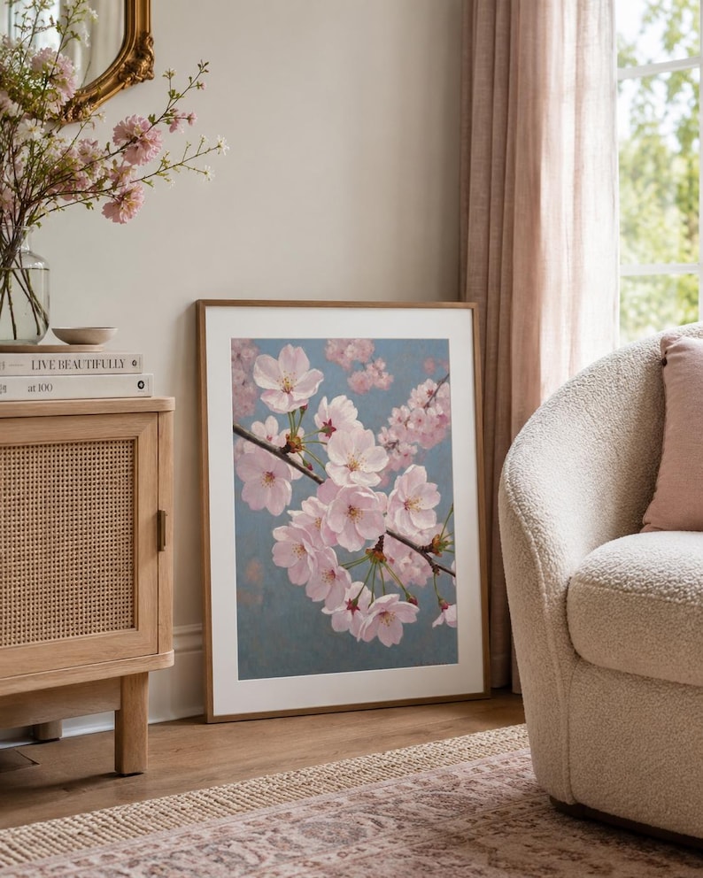 以下が含まれることがあります： 淡いピンク色の桜の花が咲く枝が描かれた、青色の背景の額入りアートプリント。木製キャビネットとクリーム色の肘掛け椅子が置かれたリビングルームに飾られています。プリントは木製のフレームに入っています。