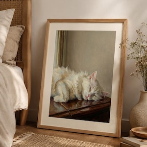 May include: A framed print of a sleeping white cat with fluffy fur. The cat is resting on a dark wooden surface. The artwork is in a light wooden frame, leaning against a wall near a bed and a woven rug. The cat's eyes are closed.