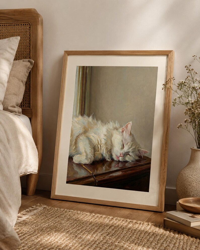 May include: A framed print of a sleeping white cat with fluffy fur. The cat is resting on a dark wooden surface. The artwork is in a light wooden frame, leaning against a wall near a bed and a woven rug. The cat's eyes are closed.