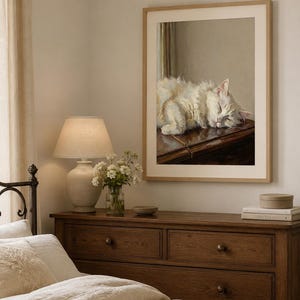 May include: A framed print depicts a sleeping white cat on a dark wooden surface. The print hangs above a wooden chest of drawers, a lamp, flowers, and books. The room has a neutral colour palette and soft lighting.