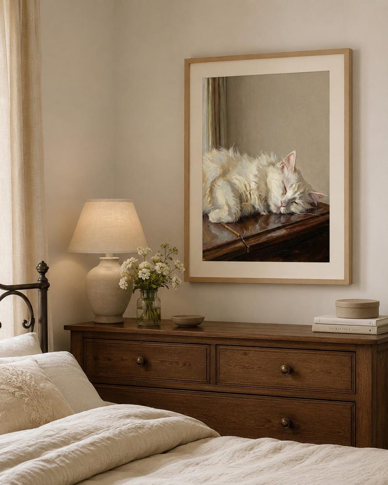 May include: A framed print depicts a sleeping white cat on a dark wooden surface. The print hangs above a wooden chest of drawers, a lamp, flowers, and books. The room has a neutral colour palette and soft lighting.
