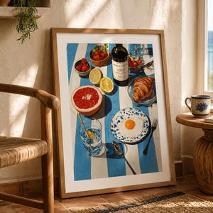 May include: A framed print depicting a breakfast scene on a blue and white striped tablecloth. The image features a bottle of wine, a croissant, a plate with a fried egg, fruit, and a glass of water. A wooden chair and a small table with a mug are in the foreground.