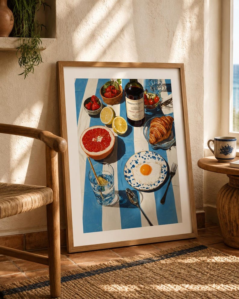 May include: A framed print depicting a breakfast scene on a blue and white striped tablecloth. The image features a bottle of wine, a croissant, a plate with a fried egg, fruit, and a glass of water. A wooden chair and a small table with a mug are in the foreground.