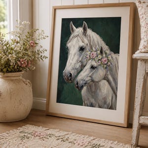 Op de afbeelding: Een ingelijste kunstdruk met twee witte paarden, waarvan &eacute;&eacute;n versierd met een bloemenkrans. Het kunstwerk staat tegen een donkergroene achtergrond en is ingelijst in licht hout. De print leunt tegen een muur in de buurt van een vaas met bloemen en een patroonkleed.