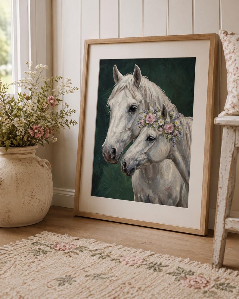 Op de afbeelding: Een ingelijste kunstdruk met twee witte paarden, waarvan &eacute;&eacute;n versierd met een bloemenkrans. Het kunstwerk staat tegen een donkergroene achtergrond en is ingelijst in licht hout. De print leunt tegen een muur in de buurt van een vaas met bloemen en een patroonkleed.