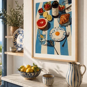 May include: A framed print depicting a breakfast scene with a blue and white striped tablecloth. The print features a bottle of wine, fruit, a croissant, and a fried egg on a plate. A bowl of lemons and a striped jug are on a marble countertop.