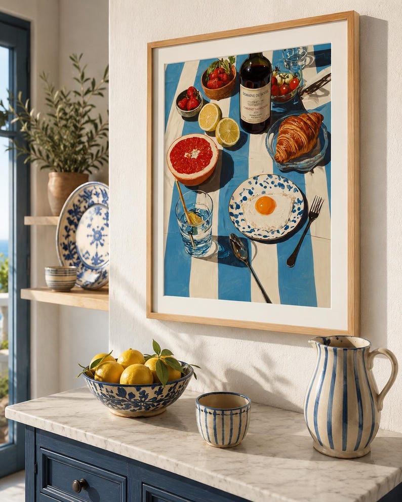 May include: A framed print depicting a breakfast scene with a blue and white striped tablecloth. The print features a bottle of wine, fruit, a croissant, and a fried egg on a plate. A bowl of lemons and a striped jug are on a marble countertop.