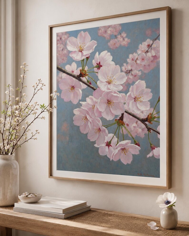 以下が含まれることがあります： 桜の花を描いた額装されたアート作品。淡いピンク色の花が枝に咲き、背景は柔らかな青色。明るい茶色の木製フレームに収められており、インテリアに最適です。