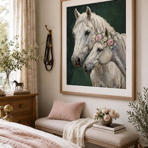 Op de afbeelding: Een ingelijste prent van twee witte paarden, waarvan &eacute;&eacute;n met een bloemenkrans, tegen een donkergroene achtergrond. Het kunstwerk is in een lichte houten lijst. De kamer bevat een bank, een roze kussen en een vaas met bloemen.