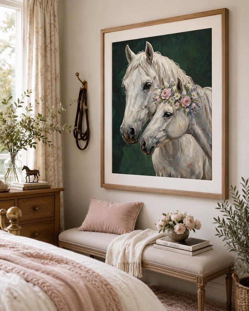 Op de afbeelding: Een ingelijste prent van twee witte paarden, waarvan &eacute;&eacute;n met een bloemenkrans, tegen een donkergroene achtergrond. Het kunstwerk is in een lichte houten lijst. De kamer bevat een bank, een roze kussen en een vaas met bloemen.