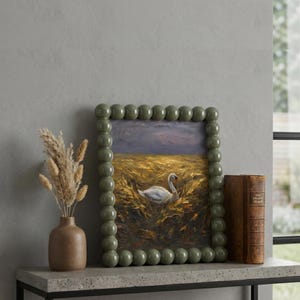 May include: A framed oil painting featuring a swan in a field, with a green beaded frame. The painting sits on a grey shelf, accompanied by a brown vase with dried flowers and a vintage book. The style is rustic and elegant.
