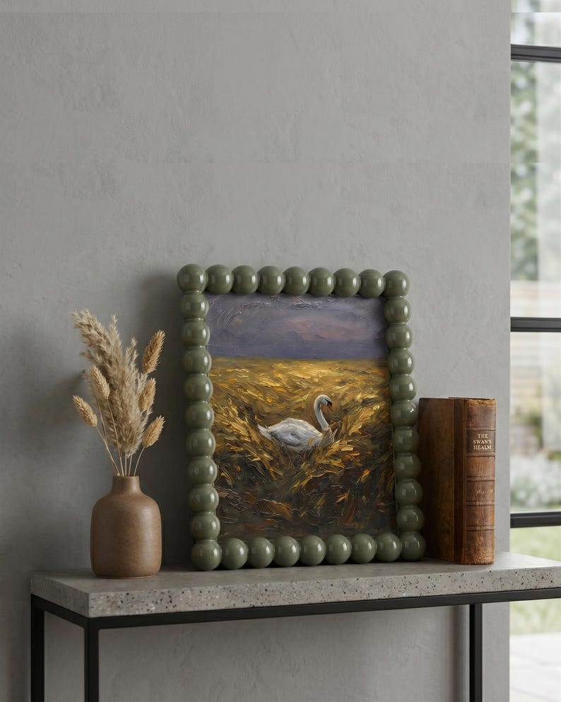 May include: A framed oil painting featuring a swan in a field, with a green beaded frame. The painting sits on a grey shelf, accompanied by a brown vase with dried flowers and a vintage book. The style is rustic and elegant.
