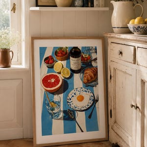 May include: A framed print depicting a breakfast setting with a blue and white striped tablecloth. The image features a bottle of wine, fruit, a croissant, and a plate with a fried egg. A glass of water and a spoon are also visible.