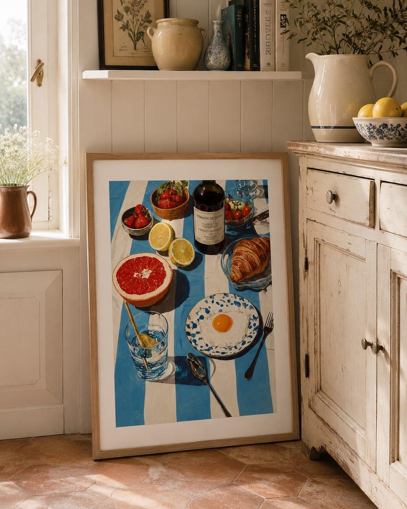May include: A framed print depicting a breakfast setting with a blue and white striped tablecloth. The image features a bottle of wine, fruit, a croissant, and a plate with a fried egg. A glass of water and a spoon are also visible.