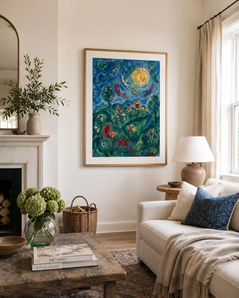 May include: A colourful framed artwork hangs above a white sofa in a living room. The artwork depicts a surreal landscape with a bright sun, figures, and flowers. A coffee table with books and a vase of flowers sits in front of the sofa.