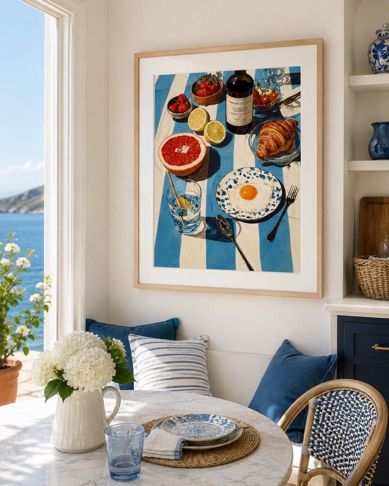 May include: A framed print shows a breakfast scene with a blue and white striped tablecloth. The image includes a grapefruit, strawberries, a croissant, a fried egg, and a bottle of wine. A glass of water and a blue and white patterned plate are also visible.