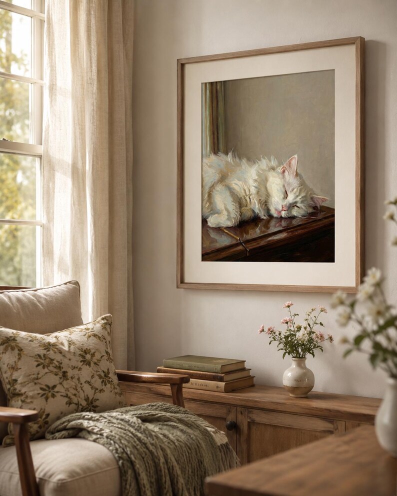 May include: A framed print depicting a sleeping white cat with pink ears. The artwork is in a light brown wooden frame. The room features a wooden cabinet, a chair with a floral pillow, and a window with sheer curtains. The image is in a room.