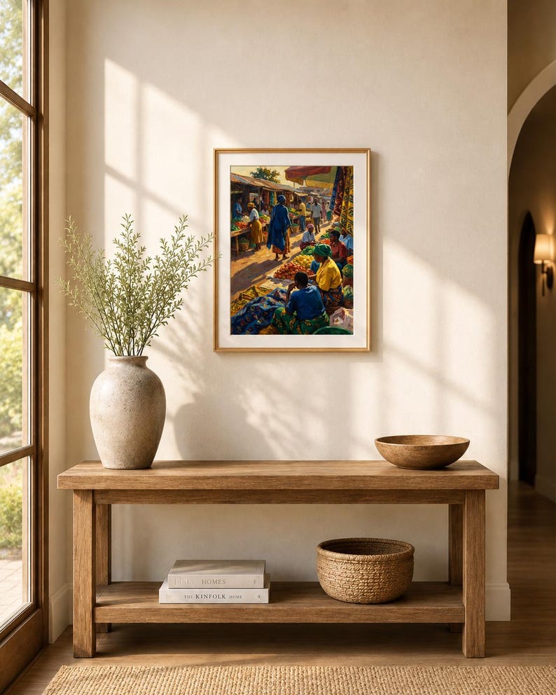 May include: A framed print showing a bustling outdoor market with figures in bright attire. The artwork hangs above a wooden console table, adorned with a vase of foliage, books, a woven basket, and a wooden bowl.