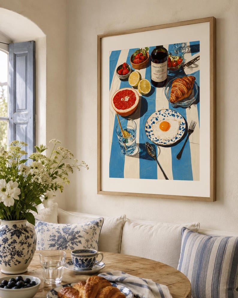 May include: A framed print depicting a breakfast scene with a blue and white striped tablecloth. The print includes a bottle of wine, a croissant, a grapefruit, strawberries, and a fried egg on a plate. A vase of white flowers and a table setting are in the foreground.