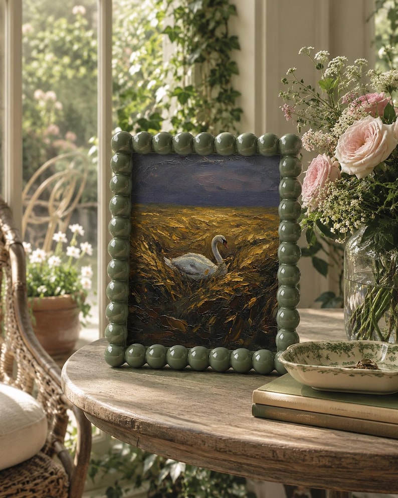 May include: A painting of a swan in a field, framed in a light green beaded frame. The painting sits on a wooden table, with a vase of pink roses and other flowers. A small dish and books are also on the table.