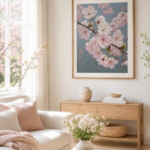以下が含まれることがあります： 淡い青色の背景に、枝に咲くピンク色の桜の花が描かれた額入りのアートプリント。プリントは薄茶色の木製フレームに入っています。この画像は、白いソファと木製家具のある明るいリビングルームに飾られています。