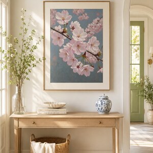 以下が含まれることがあります： 淡い青色の背景にピンク色の桜の花が描かれた額入りのアートワーク。アートワークは、編まれたバスケット、装飾的なボウル、青と白のセラミックジャーを備えた木製のコンソールテーブルの上に展示されています。