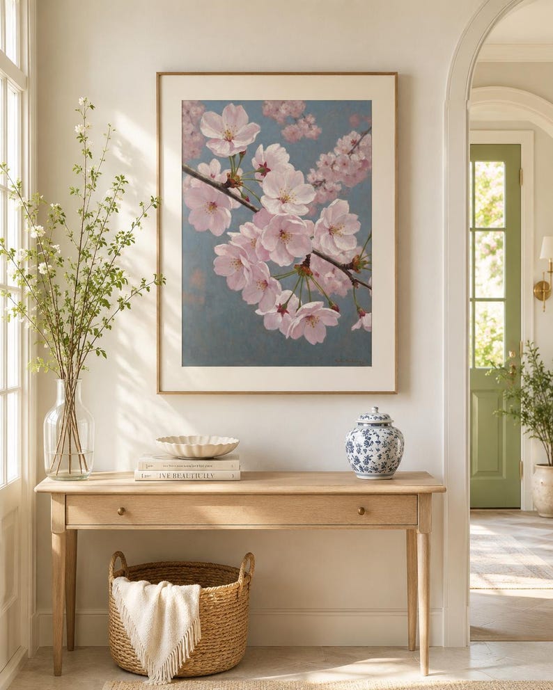 以下が含まれることがあります： 淡い青色の背景にピンク色の桜の花が描かれた額入りのアートワーク。アートワークは、編まれたバスケット、装飾的なボウル、青と白のセラミックジャーを備えた木製のコンソールテーブルの上に展示されています。