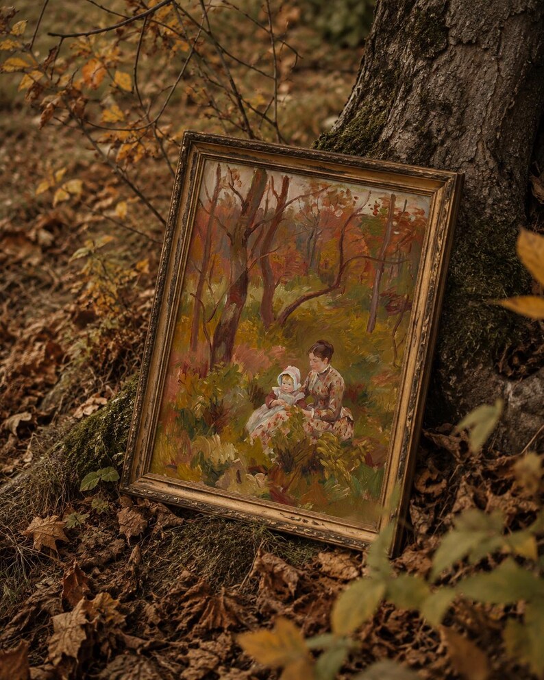 May include: An antique framed painting depicting a woman and child in a forest setting. The artwork features warm tones of brown, orange, and green, with a gold-colored frame. The painting is leaning against a tree trunk, surrounded by fallen leaves.