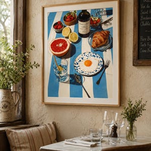 May include: A framed print depicting a breakfast setting with a blue and white striped tablecloth. The artwork includes a bottle of wine, fruit, a croissant, and a fried egg on a plate. A glass of water and a spoon are also present.