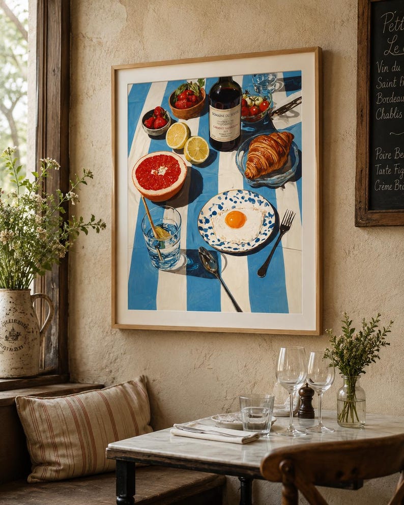 May include: A framed print depicting a breakfast setting with a blue and white striped tablecloth. The artwork includes a bottle of wine, fruit, a croissant, and a fried egg on a plate. A glass of water and a spoon are also present.