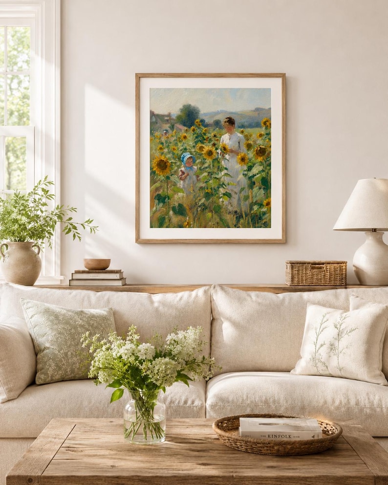 Puede incluir: Un cuadro enmarcado representa a una mujer y un ni&ntilde;o en un campo de girasoles. La obra de arte, en tonos c&aacute;lidos, se exhibe sobre un sof&aacute; beige con cojines. Una mesa de centro de madera sostiene un jarr&oacute;n con flores blancas y una bandeja tejida.
