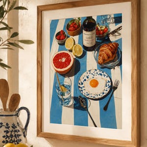 May include: A framed print depicting a breakfast setting on a blue and white striped cloth. The image features a bottle of wine, fruit, a croissant, and a fried egg on a plate. A glass of water and a spoon are also present.