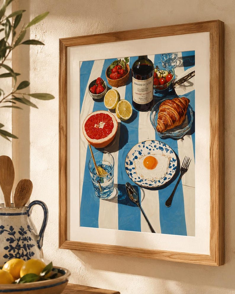 May include: A framed print depicting a breakfast setting on a blue and white striped cloth. The image features a bottle of wine, fruit, a croissant, and a fried egg on a plate. A glass of water and a spoon are also present.
