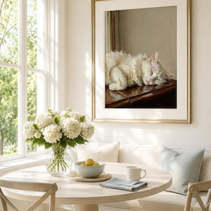 May include: A framed painting of a sleeping white cat with a gold frame hangs above a round dining table. The table is set with a vase of white flowers, a bowl of lemons, and a mug. The room has a window and a built-in bench.
