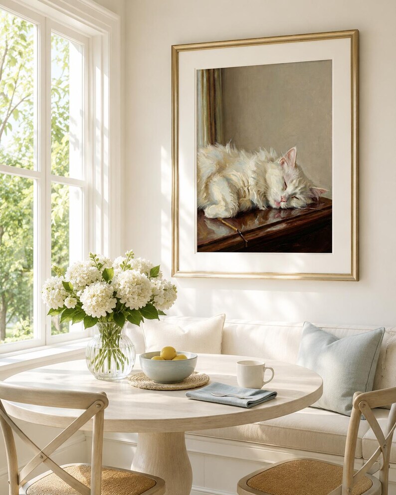 May include: A framed painting of a sleeping white cat with a gold frame hangs above a round dining table. The table is set with a vase of white flowers, a bowl of lemons, and a mug. The room has a window and a built-in bench.